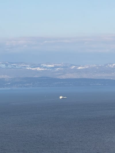 The image depicts a body of water, possibly a lake or sea, with mountains visible in the background. The mountains have patches of snow. A ship is seen on the water. The sky is light blue, suggesting a clear day. The location is most likely near Zagorje, Croatia.