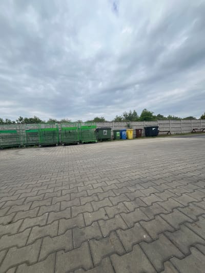 In Bogucin, Poland, a row of green metal cages on wheels stands to the left of a concrete fence. To the right of the cages, several wheeled trash bins are lined up against the fence: one dark green, one lighter green, one blue labeled "PAPIER" (paper), one yellow, one brown, and one black. The ground is paved with interlocking concrete blocks. Trees and foliage are visible behind the fence. The sky is overcast with gray clouds.