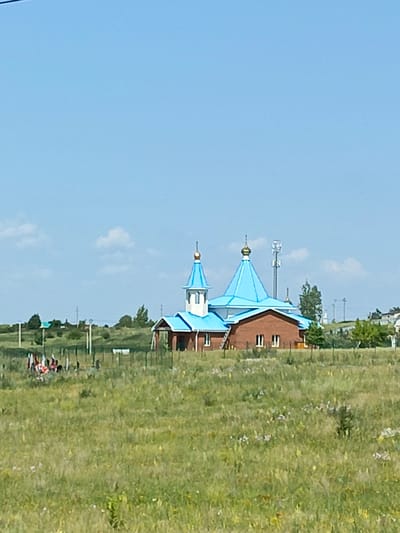 A brick church with bright blue domes stands in a grassy field under a clear blue sky in Lekarevka, Russia. A group of people are gathered in the foreground, some holding flags. The church has a traditional architectural style with multiple domes and a cross on top. In the background, trees and some power lines can be seen, suggesting a rural or semi-rural setting. The field appears to be a meadow with wildflowers.