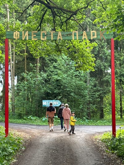 In Gorka, Russia, a path leads into a forest through a wooden archway. The arch is painted green with gold Cyrillic letters spelling "ФИЕСТА ПАРК" (Fiesta Park). Red poles support the arch. A group of people, including two adults and a child, walk away from the camera on the path, heading towards a directional sign also indicating "ФИЕСТА ПАРК." The environment is lush with dense green trees and foliage.