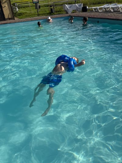 In the outdoor pool of Nechkino, Russia, a young boy is seen floating on his back, wearing blue arm floaties with a white steering wheel design and blue patterned swim trunks. His body is mostly submerged in the clear, turquoise water, with his legs and feet visible below the surface. The sunlight creates rippling patterns on the water's surface. In the background, other children are visible in the pool, and beyond that, lounge chairs and grassy areas can be seen.