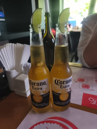 Two bottles of Corona Extra beer are prominently displayed in the foreground of this media file. Each bottle is topped with a wedge of lime, a classic garnish for the beverage. The bottles are positioned on a table, likely in an outdoor or semi-outdoor setting, suggested by the natural light and greenery visible in the background. The scene appears to be casual and relaxed, possibly a meal or a gathering.

The label on each bottle clearly reads "Corona Extra" and includes information such as "Alc. 4.5% Vol." and "LA CERVEZA MAS FINA". There is also a date stamped on one bottle: "07/11/2026". In the background, hints of other elements are visible, including what looks like a salt and pepper shaker set, a stack of white paper napkins in a holder, and the blurred figures of people, one of whom is wearing a white garment. The overall impression is one of leisure and enjoyment, with the focus squarely on the refreshing drinks. The presence of a second bottle suggests shared consumption or a choice between options. The lighting and the natural elements in the background suggest it could be daytime.