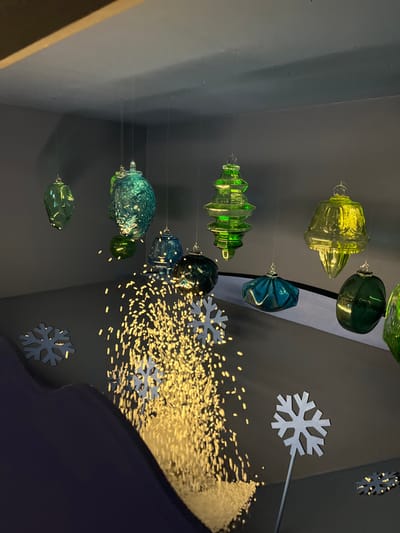 An indoor decorative display is depicted within the city of Sélestat, France. Numerous translucent glass or plastic ornaments are suspended from a dark grey ceiling by thin wires. These ornaments vary in shape, including bell-like, pinecone-like, faceted, and elongated forms, and are colored in shades of green, teal, blue-green, and yellowish-green, some exhibiting textured surfaces. Below the suspended ornaments, a cascade of small, bright, light-colored particles, resembling glitter or artificial snow, is actively falling towards the lower left, intensely illuminated. Several flat, white or light-grey snowflake cutouts are positioned on a dark grey surface below the falling particles; one larger snowflake stands upright on a slender stick in the mid-right foreground. The environment includes a dark grey ceiling and wall, with a lighter grey-beige section visible in the upper left corner. A curved, dark blue or purple structure partially obscures the lower left foreground. No individuals are present.