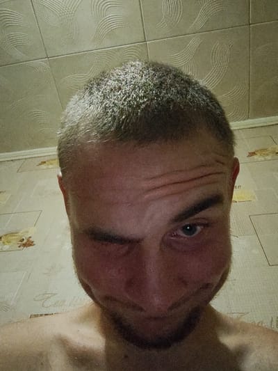 The image shows a close-up, low-angle selfie of a man with short, light brown hair. He is looking directly at the camera with his right eye open and his left eye squinted shut. His forehead is furrowed, creating horizontal lines above his eyebrows. He has a faint stubble on his chin and jawline. His skin is flushed, particularly on his cheeks. He appears to be shirtless, with his shoulders and upper chest visible.
The background consists of tiled walls and a tiled floor. The wall tiles are a light, beige color with a textured pattern of curved lines. The floor tiles are also a light color, possibly beige or cream, with a repeating pattern of floral or abstract motifs in a darker shade. The lighting in the image is dim and appears to be coming from above and slightly in front of the subject.
The image appears to have been taken indoors, possibly in a bathroom or a similarly tiled room. There is no specific information in the image to indicate its location within the city of Okhtyrka, Ukraine.