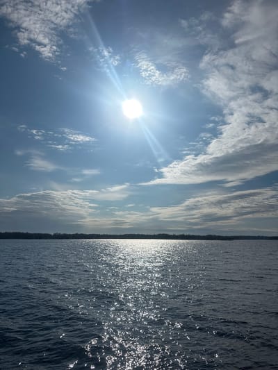 The image captures a bright, expansive outdoor scene, divided horizontally between sky and water by a distant treeline. The upper two-thirds of the frame display a vibrant blue sky dotted with scattered clouds, ranging from wispy formations to more textured patterns. The sun, a brilliant disc, is positioned on the left side, emitting strong rays and a prominent lens flare that illuminates the sky.

A narrow, dark band of land, likely a distant forest or shoreline, forms the horizon, stretching across the entire width of the image. Below this horizon, the lower third of the photograph reveals a large body of water, its surface disturbed by numerous small ripples. The intense sunlight reflects brilliantly off the water, creating a sparkling, shimmering effect with numerous white highlights and streaks across its dark surface.

The scene depicts a sunny day, likely midday or early afternoon given the sun's high intensity. The weather appears clear with some cloud cover. The presence of a wide body of water with a distant treeline, coupled with the context, suggests a location like the Daugava River near Ikšķile, Latvia. No people, man-made objects, or discernible text are visible in the image. The overall mood is one of natural brightness and serenity.