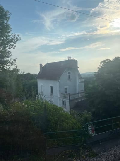 An exterior, medium shot captures a pale blue, multi-story house with a red roof, nestled amidst lush green trees and foliage. The house features several windows, some with arched tops, and a balcony with ornate railings. A chimney rises from the roof. The sky above is a mix of blue with wispy white clouds, illuminated by the soft glow of the sun peeking through. In the foreground, a dark green metal fence runs horizontally across the lower portion of the image, with a gravel path visible below it. The scene appears to be a residential area in Sartrouville, France, offering a glimpse into a tranquil, verdant setting.