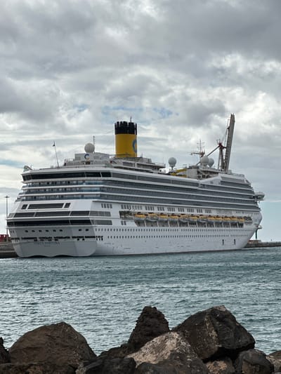 A large, white multi-deck cruise ship named "COSTA FORTUNA," homeported in Genoa, Italy, is docked in the port of Puerto del Rosario, Fuerteventura, Spain. The vessel features a prominent yellow and black funnel with a blue "C" logo on its upper deck, alongside multiple white radar domes, various antennas, and a large crane structure towards the stern. Numerous yellow lifeboats are stowed along the ship's starboard side, below rows of windows and balconies. The ship is positioned on choppy, dark blue-green water. In the immediate foreground, dark, irregular volcanic rock formations are partially submerged, indicating a rocky shoreline or breakwater. The sky above is entirely overcast with dense grey clouds. No individuals are visible within the frame, and the ship appears stationary.