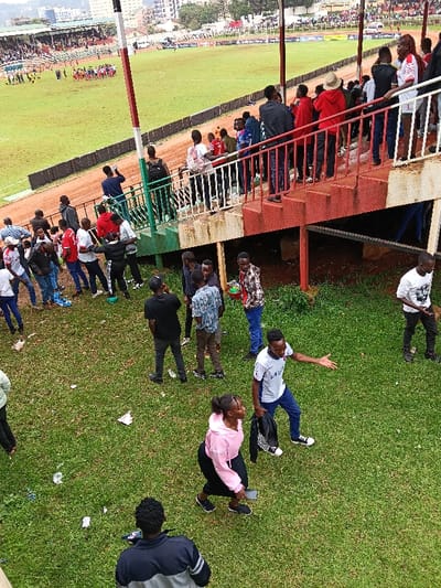 The image captures a scene at Gusii Stadium in Kisii Central ward, Kisii County, Kenya. The foreground consists of a sloped, grassy area extensively littered with paper and plastic debris. Approximately fifteen individuals, predominantly young adults, are present on this grass, either standing or walking. Notable figures include a woman in a pink long-sleeved top and dark skirt, holding a mobile phone in her left hand while walking right. Adjacent to her, a man in a white and grey striped T-shirt and blue trousers gestures with his right arm, holding a black jacket in his left hand. Several other men in casual attire are grouped, engaged in conversations.
In the midground, a multi-tiered red concrete stairwell with white railings ascends towards the upper stadium levels. Numerous spectators, mainly young men, are standing on these stairs and an adjacent lower green-painted concrete platform. A prominent red and white striped pole extends vertically from this structure.
The background reveals the stadium's large green football pitch, partially worn, bordered by a reddish-brown dirt track. A cluster of individuals, possibly players or officials, in yellow and red uniforms, is visible near the center of the pitch, with other small groups dispersed. Beyond the track, multi-tiered stadium stands are packed with spectators. The distant horizon shows urban buildings and hills under an overcast sky.
