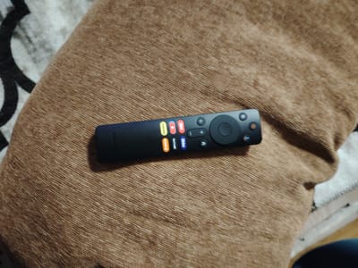 The media file is a close-up, slightly angled shot of a black TV remote control resting on a textured brown fabric surface, likely a cushion or blanket. The remote is the primary subject, positioned horizontally across the frame. It features several colored buttons with icons and text, including what appear to be app shortcuts like "YouTube", "Prime Video", and "Netflix," indicated by their respective logos. There's also a directional pad with a central "OK" button, volume controls, and a power button. The fabric background has a distinct ribbed texture and a warm, earthy tone. The lighting suggests an indoor setting, possibly during the daytime, with no strong shadows indicating a specific time. No people are visible. The overall impression is a simple, everyday scene, focusing on the technology of the remote control. The context provided indicates the location as Gorshki, Russia, but there are no specific visual cues within the image to confirm this. The visible text on the remote includes app names and potentially brand markings, but these are not clearly legible in the provided image.