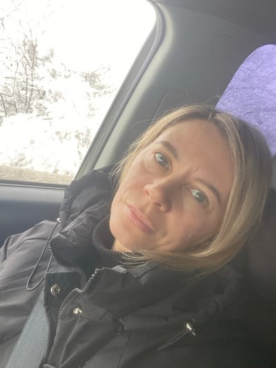 An adult female, characterized by light-colored hair and an unsmiling expression, is positioned in the passenger seat of a vehicle in Abinsk, Russia. She wears a black puffer jacket over a black turtleneck, with a gray seatbelt strap visible across her chest. Her gaze is directed towards the camera, suggesting a self-portrait. Through the left side window, a winter landscape is visible, featuring snow-covered trees and a bright, overcast sky. The right side window displays a muted purple tone, potentially due to tinting or reflection. The vehicle's interior trim, including part of the headliner and window frame, is visible and appears to be a dark gray color.