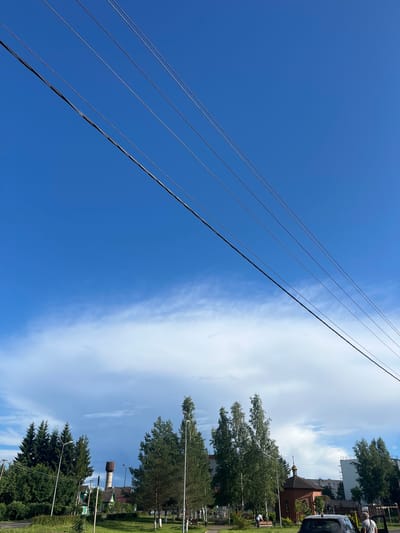 The image, taken in the city of Serebryansky, Russia, depicts a scene under a bright blue sky with thin white clouds. Four dark power lines stretch diagonally across the upper portion of the frame. The middle ground features a collection of deciduous trees with green foliage and a few evergreen trees on the left. In the distance, a water tower stands tall, and a small, brick chapel with a golden dome is visible. Several streetlights are scattered throughout the area. A dark-colored car is parked in the foreground on the right, with its rear door open and a person standing beside it. Another person is seen seated on a bench to the left of the chapel. The overall environment suggests a sunny day in a semi-urban or suburban setting.