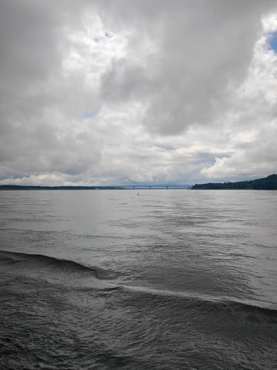 The image depicts a wide river under a cloudy sky. In the distance, a long bridge spans the water, connecting the two shores. The riverbanks are lined with dense green trees. The water in the foreground is dark and choppy, with visible ripples and waves. The sky is filled with heavy, grey clouds, with only small patches of blue sky visible. The scene appears to be a natural landscape with man-made structures. The location is specified as Vysokogorsky District, Russia.
