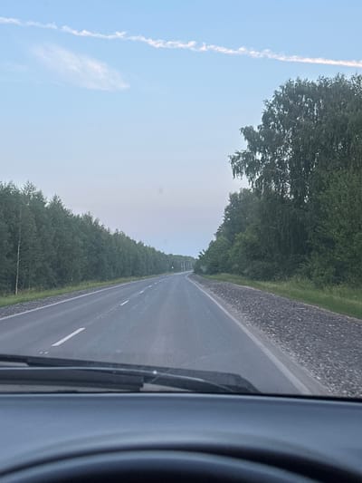 The image is a point-of-view shot from inside a vehicle, looking out at a two-lane asphalt road with white lane markings. The road is flanked by dense green trees on both sides, forming a forest. The sky is a pale blue with a few wispy white clouds. The scene appears to be a highway in a rural or semi-rural area, possibly on the outskirts of Pervomayskiy, Russia, given the context. The lighting suggests it is either early morning or late afternoon. The foreground shows a portion of the vehicle's dashboard and windshield wipers. There are no people or other vehicles visible in the frame.