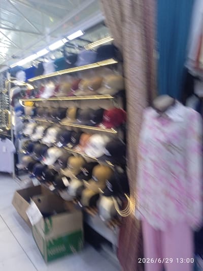 The image depicts an indoor market stall, likely in Ak-Suu, Kyrgyzstan, filled with numerous baseball caps displayed on shelves. The lighting is bright, with fluorescent lights visible overhead. To the right of the caps, a mannequin is dressed in a pink and white patterned shirt and pink pants, suggesting a clothing section within the market. In the foreground, a cardboard box is visible. The overall scene is somewhat blurry, with motion blur indicating movement during the photograph. The timestamp "2026/6/29 13:00" is superimposed on the bottom right corner of the image.