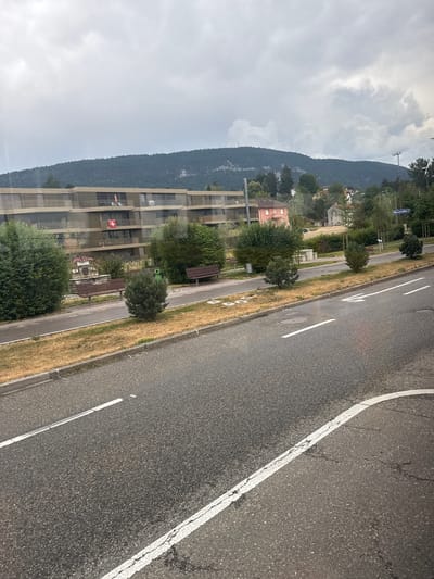 The image captures a street scene in Reconvilier, Switzerland. The foreground is dominated by an asphalt road with white lane markings, showing signs of wear and tear. A curb and a dry, grassy verge separate the road from a paved pedestrian walkway. Along the walkway, there are two dark-colored benches and a small, conifer tree. Behind the walkway, a multi-story building with balconies and a light-colored facade is visible. A Swiss flag is displayed on one of the balconies. Further in the background, more buildings and lush green trees are scattered across a hillside, which eventually merges into a densely forested mountain range under a cloudy sky. A street sign labeled "Reconvilier" is positioned on the right side of the image, indicating the location. The overall impression is that of a residential area bordering a natural landscape on an overcast day.