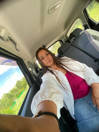 A young woman with long, brown hair is sitting in the back seat of a car, taking a selfie. She is wearing a pink tank top and an open white shirt. Her left arm is extended towards the camera, and she is wearing a black bracelet with a star-shaped charm. The car's interior is visible, including the headrests and seats. Outside the car window, a green, grassy landscape with trees and a blue sky with clouds can be seen. The specific location in the city of Yuldybayevo, Russia, is not discernible from the image.