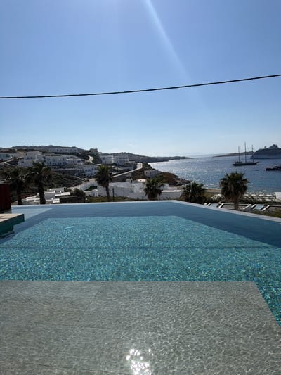 The image shows a view from what appears to be an infinity pool overlooking the town and harbor of Mykonos, Greece. The town is characterized by white-washed buildings that are clustered together on the hillside. The Aegean Sea is in the background with a cruise ship and several sailboats visible on the horizon. Palm trees are interspersed among the buildings, contributing to the scenery. A black power line is positioned in the left of the sky. The pool is lined with a blue mosaic finish, and the deck area adjacent to the pool is paved with gray tiles.