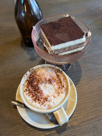 The scene depicts a dessert and beverage arranged on a wooden table in an indoor setting within Dorchester, United Kingdom. A square slice of tiramisu, layered with biscuit and cream and dusted with cocoa powder, rests in a clear pink, ribbed glass dessert bowl with a stem, positioned towards the top-right of the frame. A small portion of the tiramisu's upper right edge appears dislodged. Below and to the left of the tiramisu, a light-colored ceramic cup filled with a frothy beverage, likely a cappuccino or latte, is visible. The beverage's foam is generously topped with cocoa powder. The cup sits on a matching saucer. A metallic spoon lies on the saucer, partially beneath the cup's handle, and a light-colored, rectangular biscuit is placed on the saucer next to the cup. In the blurred background, an amber-colored glass bottle stands upright. The wooden table surface exhibits a distinct grain pattern. No people are visible.