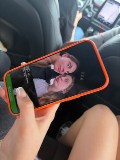A close-up image displays a person holding an orange-cased smartphone, which displays a photograph of two young women making silly faces. The metadata on the phone screen indicates "aleksandra," "Sun May 31 2020," "15:25," "Aleksandarovo, Bulgaria," and coordinates "42.9808, 25.3890." The background shows the interior of a car, specifically the dashboard and passenger seating area. The person holding the phone has their thumb near the bottom of the screen, seemingly about to press a button that says "Memory Uploaded." The environment suggests they are sitting in a car, possibly within the city of Chokoba, Bulgaria.