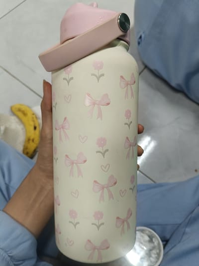In a residential area of Cileungsi, Indonesia, a hand holds a white water bottle adorned with pink bows and flowers. The water bottle has a pink lid. To the left, a yellow banana rests on a white cloth. The scene is illuminated by artificial light, creating reflections on the water bottle and a metallic object on the floor.