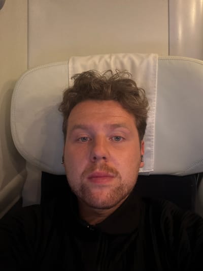 A man is lying down on a white cushioned seat. He has light skin, curly brown hair, and a beard. He is wearing a black top. The location is in Milton Keynes, United Kingdom.
