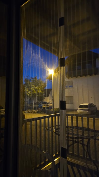 The image depicts a nighttime view through a screened window or balcony door in Vöhringen, Germany. The foreground is dominated by the textured screen and its vertical supports. Beyond the screen, a street lamp illuminates a residential area. A tree with green foliage stands prominently, with houses and vehicles visible in the background. A white station wagon is parked on the right side of the street. The sky above is a deep blue, suggesting twilight or early night. A metal railing and outdoor furniture are visible on a patio or balcony in the lower part of the image. The overall scene suggests a quiet evening in a suburban or small-town setting.