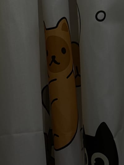 The image shows a close-up of a white fabric curtain with cartoon cat patterns. One cat is yellow with a black nose and eyes, wearing a brown costume with a black outline. Another cat is black with white eyes and is partially visible on the right side. The curtain is rolled or folded, creating a shadow and highlighting the texture of the fabric. There is a dark circular shape on a white background on the upper right of the image, possibly part of another cat's face or a design element. The image is dimly lit, with soft shadows. The location is the city of Poritsy, Russia.