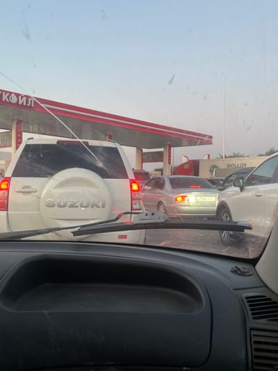 The image shows a traffic jam at a gas station in Troitsk, Russia. The scene is viewed from inside a vehicle, looking forward. In the foreground, the dashboard of the vehicle is visible. Beyond the dashboard, several cars are lined up, waiting to refuel. A white Suzuki SUV is prominently visible, with its rear facing the camera. To the right of the SUV, a silver sedan with a Russian license plate (A739ET 16) is also in the queue. In the background, the red canopy of the gas station is visible, with signage that includes the word "КОЙЛ" and numbers indicating fuel prices. Further back, a white truck with the word "MOLLOY" written on its side is parked. The sky is overcast with a light blue hue, suggesting it may be early morning or late afternoon. The overall impression is one of a typical traffic scene at a busy fuel station.