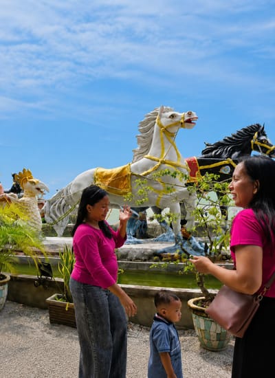 The image depicts an outdoor scene at what appears to be a tourist attraction in Kota Pari, Indonesia. In the foreground, two women in vibrant pink tops are interacting, one looking towards the other and gesturing with her hand. A young child in a blue shirt stands between them, looking off to the side. Behind the people are large, ornate statues of horses, one white with a golden saddle and harness, and another black. To the left, a statue of a mythical creature resembling a bird with a crown is visible. The statues are positioned near a body of water, likely a pond or pool, surrounded by potted plants. The background features a clear blue sky with scattered clouds. The ground is covered in gravel or small stones. The overall atmosphere suggests a sunny day at a decorative outdoor location.
