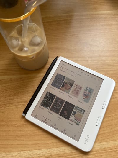 The image shows a Kobo e-reader placed on a light brown wooden surface, next to a tall glass of iced coffee. The e-reader displays a screen with multiple book covers, indicating a home screen with a list of available books. A black stylus pen lies diagonally across the e-reader. The glass of iced coffee contains ice cubes and a straw. The wooden surface appears to be a table. There is nothing in the image to indicate the location in High Wycombe.
