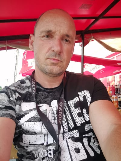 A middle-aged bald man with light skin and stubble is taking a selfie under a red canopy. He is wearing a black t-shirt with a white graphic design and a black lanyard around his neck. The background shows a shelf of bottles, possibly drinks, and part of a red umbrella. The image appears to have been taken outdoors, potentially at a market or outdoor event. The man is looking directly at the camera with a neutral expression. The image does not provide any specific location information within Dralfa, Bulgaria.