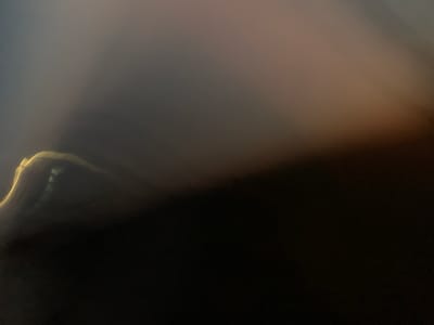The image is an abstract, blurred, low-light photograph. It features a large expanse of darkness in the lower right portion of the frame, transitioning into a lighter, smoky or foggy area in the upper left. A curved, luminous line, possibly representing a light source or a reflective edge, is visible in the bottom left quadrant, cutting across the darkness. The overall impression is one of indistinct shapes and muted colors, dominated by dark tones with a gradient of grays and hints of warm, light brown or orange in the upper right. The image does not contain any identifiable landmarks, people, or specific objects that would allow for contextualization within Uzunovo, Russia, or any other location.