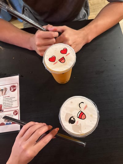 The image depicts a high-angle view of a dark, matte-finish table within a bubble tea shop located in North Miami Beach, United States of America. Two clear plastic cups containing beverages are centrally positioned. The upper cup holds an orange-yellow liquid with dark spherical inclusions, sealed with a plastic film displaying a cartoon face featuring two red heart-shaped eyes, pink blush marks, and an open mouth. The lower cup holds a lighter, opaque liquid, also with dark spherical inclusions, sealed with a plastic film displaying a winking cartoon face with one eye closed, an open mouth showing teeth, and pink blush marks.
In the upper portion of the frame, the forearms and hands of one individual are visible, wearing a dark grey shirt over a light blue layer. Their hands are loosely clasped, resting on the table, and they are holding a metallic reusable straw. In the lower portion of the frame, the left hand of a second individual is visible. This hand has fingernails painted with orange/red tips and holds a black plastic straw, partially encased in its plastic wrapping, positioned near the lower beverage.
A laminated menu is partially visible on the left side of the table. It features red headings and lists "Fruit tea" options such as "Strawberry black tea," "Peach black tea," and "Passion Fruit tea," with a price of "$6.50." Another section for "Popping boba" is partially visible with a price of "$6.75."