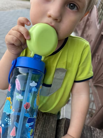 A young child, wearing a bright yellow t-shirt with a dark trim, is focused on drinking from a blue water bottle adorned with cartoon dinosaurs and other colorful stickers. The child's face is partially visible, with one eye looking towards the camera. The child's left hand is holding a green sippy cup lid, which is attached to the water bottle. The background appears to be outdoors, with hints of wooden furniture, possibly a picnic table, and a blurred background that suggests a park or garden setting. The lighting suggests it is daytime. There is no visible text. The overall scene conveys a casual, everyday moment of a child enjoying a drink.