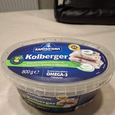 The image shows a plastic tub of "KOLBERGER" brand Matjes herring in plant oil with marinated onions. The tub is predominantly blue with white lettering and features an illustration of herring fillets, onions, and parsley. It also includes a DLG quality seal and nutritional information indicating a high omega-3 content. The tub is placed on a light-colored, textured surface, likely a table. In the background, a dark, puffy object, possibly a bag or jacket, is visible. The lighting suggests an indoor environment. No specific location within the city of Bösel, Germany is identifiable from the image.