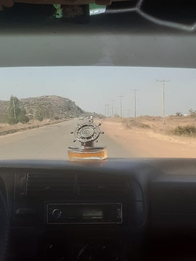 The image displays a forward-facing view from inside a vehicle, located on a paved road in Barakin Columbai, Nigeria. The foreground shows the vehicle's dark-colored dashboard, which includes a car stereo unit with a display and various controls, and several air vents. Centrally placed on the dashboard is a decorative object consisting of a ship's wheel or compass design atop a rectangular glass bottle. Beyond the windshield, a multi-lane asphalt road recedes into the distance. The road is bordered by dry, light brown soil and sparse vegetation. To the left, a prominent rocky hill with scattered trees and scrub brush rises. To the right, a series of utility poles with crossarms line the roadside, extending towards the horizon. A few distant vehicles are visible on the road. The sky above is hazy and light-colored. No identifiable individuals or specific human actions are discernible. The overall environment appears arid or semi-arid.