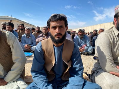 The image displays an outdoor gathering of approximately two dozen adult males seated on bare earth under a clear blue sky in Daman, Afghanistan. A young adult male, exhibiting dark hair and a full beard, occupies the central foreground, wearing a blue long-sleeved tunic (shalwar kameez), a light brown vest, and a white patterned skullcap. He looks directly forward towards the viewer. Surrounding him, other men, predominantly adults, are seated in similar positions, dressed in traditional Afghan attire, including varying colored shalwar kameez, turbans, and skullcaps. In the background, a light-colored building with a flat roof is visible to the left. A tall, light-colored concrete wall extends across the background from the center-right. A small blue plastic object rests on the ground between seated individuals in the left-midground. The scene is brightly illuminated by natural daylight, casting distinct shadows.