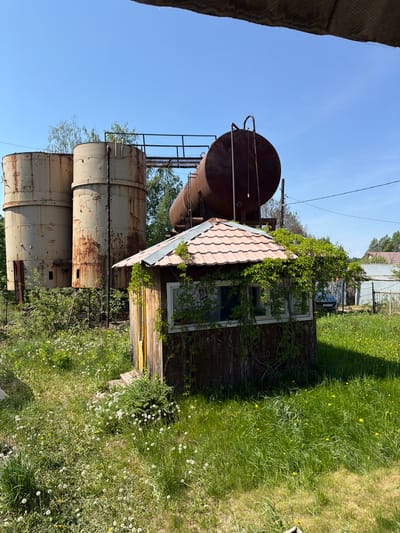 The image shows a dilapidated structure located in Ягул, Russia. Three large, cylindrical, rusty metal tanks stand vertically, topped with a larger horizontal tank. A small, weathered wooden building is situated nearby, partially overgrown with vegetation. A rusted, red-tiled roof covers part of the building. The surrounding environment consists of overgrown grass and weeds, with a glimpse of other buildings and a chain link fence in the background. The sky is clear and blue. No people are visible.