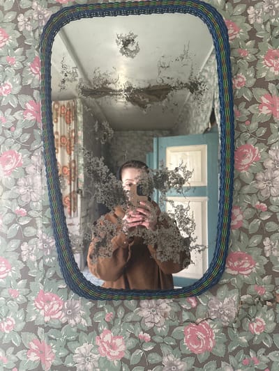 The image is a photograph taken of a person's reflection in an old, dirty mirror. The mirror has a blue wicker frame and is mounted on a wall covered in floral wallpaper. The reflection shows a person holding a smartphone, presumably taking the photo. The mirror is significantly deteriorated, with a layer of grime obscuring parts of the reflection. The background visible through the mirror shows a glimpse of a room with light blue walls and what appears to be a cabinet or shelving unit. The scene appears to be indoors, likely in a residential building in Студзёнкаўскі сельскі Савет, Belarus, given the location provided. There is a visible crack in the wall with wallpaper.