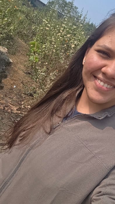 A smiling woman poses outdoors. Her brown hair falls along the left side of her body, clad in a light brown jacket with a visible zipper. Vegetation and a hint of the sky are visible in the background. The ground appears rocky and dry.