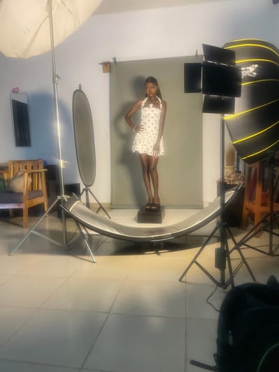 The image depicts a studio photography setup in South Ropp, Nigeria. The main subject is a young Black woman posing on a dark rectangular box, positioned in front of a grey backdrop. She is wearing a white polka dot dress and is looking towards the camera with a neutral expression. Her body is slightly contorted, with one leg bent and the other extended.
Surrounding the subject are various professional photography lights and equipment. To the left, a large, soft, umbrella-shaped light fixture is visible. Above and behind the woman, a large octagonal softbox with yellow accents is positioned, likely providing diffused lighting. Other lighting modifiers, such as black panels, are attached to stands. A curved reflector or light bouncer is positioned in front of the woman, enhancing the lighting.
The setting appears to be an indoor studio, with tiled flooring visible at the bottom of the frame. Wooden chairs and other studio furniture are present in the background. The overall lighting suggests a controlled studio environment, rather than natural daylight. The image is vertically oriented, likely due to the camera's positioning or subsequent editing. No visible text is present in the image. The activity is a staged photo shoot.