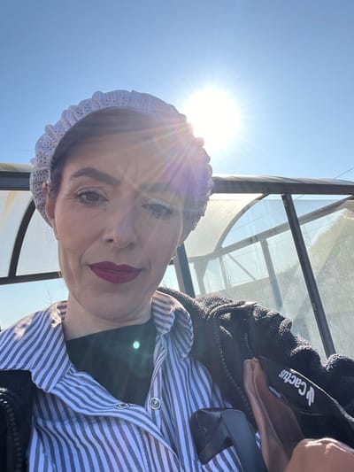 A mid-chest up, frontal portrait depicts an adult female outdoors under a clear blue sky. She wears a white, perforated, ruffled headband over dark hair, a light-colored vertically striped collared shirt over a black undershirt, and a dark, possibly fleece, jacket with a visible "Cactus" brand label. Her face shows defined eyebrows, light-colored eyeshadow, and dark red lipstick. A dark handle or armrest is partially visible in the lower right foreground. The background features a large, translucent, curved structure with dark framing, resembling a greenhouse or a shelter. The sun is prominent in the upper right sky, producing significant lens flare. The scene is located in Roodt-sur-Syre, Luxembourg.
