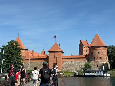 The image shows a bright, sunny day at Trakai Island Castle in Trakai, Lithuania. A group of people are walking across a wooden bridge towards the castle. The castle is a large, red brick structure with multiple towers and red-tiled roofs. In the foreground, a man in a black t-shirt with a graphic on it is walking away from the camera. To his right, a woman wearing a black tank top and shorts is walking. Behind them, several other people are also walking on the bridge. A white boat with a black roof is docked on the right side of the image, near the castle. The water is calm, and trees are visible behind the castle and on the left side of the bridge. The sky is clear and blue.