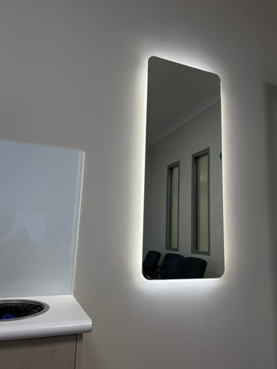 A lighted, rectangular mirror with rounded corners hangs on a white wall. The mirror reflects two rectangular skylights and a dark chair. Below the mirror is a section of a white and gray cabinet, the top of which is white and glossy. The wall is painted in a soft, light color, possibly white or light gray. The lighting around the mirror casts a bright glow.