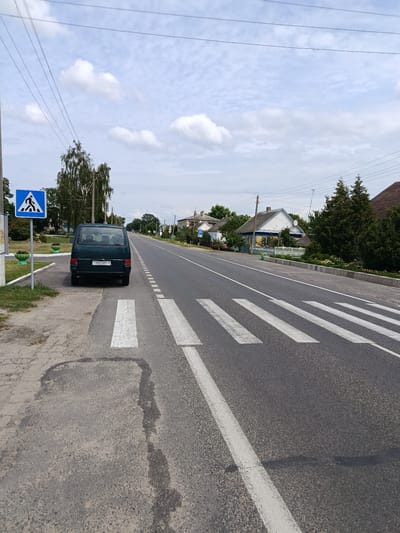 A rural road in Сошненскі сельскі Савет, Belarus, features a crosswalk and a parked dark green minivan. The minivan, with license plate "3932MT-1", is positioned on the shoulder of the road, adjacent to the crosswalk. A pedestrian crossing sign is visible to the left of the minivan. The road stretches into the distance, lined with houses and trees on both sides. The sky is partly cloudy. The overall scene depicts a quiet village setting.