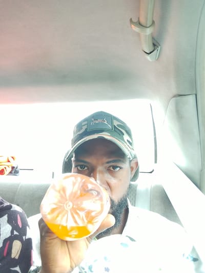 The image shows a man sitting in a car, holding a plastic bottle filled with an orange beverage. He is wearing a camouflage baseball cap with a patch on the front depicting a dog-like animal. The man has a beard and is looking at the camera. The setting appears to be the interior of a car, with the headliner and a seat visible. The bright light suggests it is daytime. A stack of folded clothing is visible in the background on the left. The man's hand is holding the bottle to his mouth, indicating he is drinking. There is no visible text in the image. The location context is Alagbede, Nigeria.