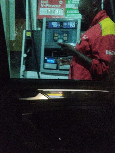 This image depicts a nighttime scene at a Shell fuel station in Kalimoni ward, Kenya, viewed from inside a vehicle. A dark-skinned individual, presumably a gas station attendant, is positioned to the right of a fuel dispenser. The attendant wears a red uniform with yellow accents on the shoulders and upper arms, featuring a "Shell HELIX" logo on the right sleeve. The individual's left hand is extended towards the central console of the fuel dispenser, appearing to interact with a small handheld device or keypad.

The fuel dispenser is gray and black with multiple digital displays. One prominent display shows "400.00" and "5.493," likely indicating a monetary value and dispensed volume. Below these figures, a smaller screen emits a blue light. Above the dispenser, a red sign with "Shell V-Power" is clearly visible, along with blurred green signage to its right. The foreground shows the dark interior of a vehicle, specifically the upper section of a door panel with a metallic handle, confirming the vantage point of the photographer. The scene is illuminated by artificial lights, and the image exhibits significant digital noise.