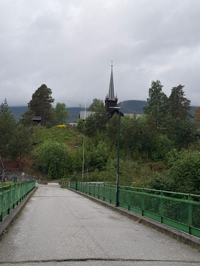 This image, taken in Vinstra, Norway, depicts a rural or semi-rural landscape dominated by a road or bridge that curves gently to the right. A green metal fence or railing runs along the left side of the asphalt road, which appears somewhat damp. On the left, beyond the railing and a dark street lamp, dense green trees and bushes cover a hillside. Through the foliage, the distinctive dark spire of a church, topped with a weather vane, is visible. Another building with a dark roof can be glimpsed further back among the trees.
In the midground, a patch of vibrant yellow vegetation adds a touch of color to the otherwise green landscape. Behind the immediate greenery, forested hills or mountains extend into the distance, fading slightly under the overcast sky. The sky is uniformly light grey and cloudy, suggesting a cool, possibly humid or post-rainy day. The overall mood is tranquil and serene, with no human activity visible. The time of day appears to be daytime, likely late morning or afternoon, given the diffuse light. Further down the road/bridge, black and yellow striped barriers are visible. No legible text is present in the image.