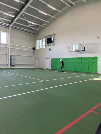 This image depicts an indoor sports hall located in the city of Gorna Mitropolia, Bulgaria. The facility features a dark green court with prominent white and red lines, delineating areas for tennis and potentially other court sports. A single adult male is positioned on the court, holding a tennis racket with the head oriented towards the ground. He is dressed in a white baseball cap, a dark short-sleeved shirt, dark patterned shorts, and athletic shoes. A knee brace is visible on his right knee. Two yellow tennis balls are present on the court: one near his right foot and another further into the mid-court area, to the left of the center line.
The hall has high, light-colored walls and a sloped ceiling with exposed white metal trusses and multiple linear fluorescent light fixtures. Natural light enters through large rectangular windows on the left wall and smaller elevated windows on the far wall. A large black flat-screen monitor, enclosed within a protective net, is mounted high on the far wall. Below it, two white doors are visible. To the right of the doors, a basketball hoop with a transparent backboard is installed. Below the basketball hoop and extending across the far wall, a series of light green padded panels are affixed. A ventilation duct runs along the upper left wall, where a black speaker is also mounted. A large grey metal utility or storage unit is visible against the lower section of the left wall.