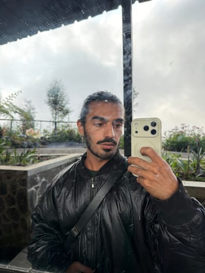 A man with dark hair tied back in a bun and a mustache takes a mirror selfie in a reflective surface. He is wearing a black, shiny bomber jacket over a green shirt and a black strap is visible across his chest. He is holding a light-colored smartphone in his right hand. The reflection shows a background with trees, plants, and a cloudy sky. The scene appears to be outdoors, possibly in a garden or park setting, with a tiled or stone border in the foreground. The location is identified as Ngadas, Indonesia.